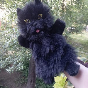 Puede incluir: Un juguete de peluche de gato negro con pelo sintético largo y desgreñado, orejas puntiagudas y grandes ojos amarillos reflectantes. El gato tiene una pequeña lengua rosa visible que sobresale de su boca y está diseñado como una marioneta.
