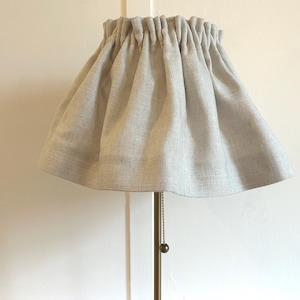 May include: A light grey lampshade with a gathered fabric design. The lampshade is made of a textured material and is attached to a brass-coloured lamp base. The shade has a flared shape and a simple, elegant design.