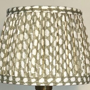 May include: A lampshade with a pleated design, featuring a repeating pattern of white and olive green geometric shapes. The shade has a scalloped edge and is designed for a lamp base. The style is vintage and decorative.