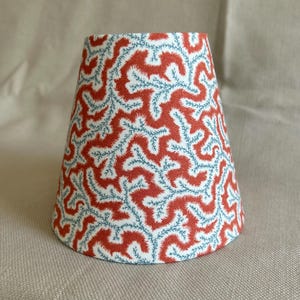 Puede incluir: Una pantalla de lámpara cónica con un patrón repetitivo rojo, azul y blanco. El diseño presenta un motivo estilizado y orgánico. La pantalla de la lámpara tiene forma de cono, adecuada para una lámpara de mesa o un accesorio similar.