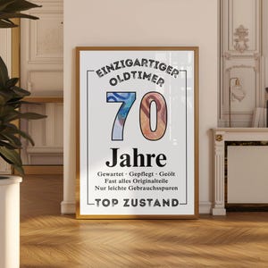 May include: Framed print with the text "EINZIGARTIGER OLDTIMER 70 Jahre" in black and white. The number 70 is in blue and orange. The print has a gold frame and is displayed against a white wall.