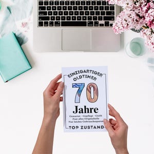 May include: A white card with the text "EINZIGARTIGER OLDTIMER 70 Jahre" is held by two hands. The card has a black border and the number 70 is in blue and brown. A laptop, flowers, and a small blue box are in the background.