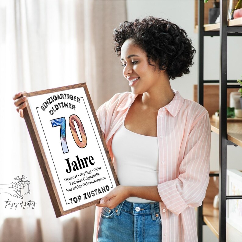 May include: A framed print with the text "EINZIGARTIGER OLDTIMER 70 Jahre" in a wooden frame. The print features the number 70 in blue and orange, with additional text below. The phrase "the joy of gifting" is in the lower left corner.