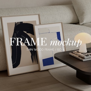 May include: Two framed abstract art prints with a white mat and a thin wood frame. The frames are on a wooden floor with a white rug and a dark wood coffee table. A white lamp with a round base is on the table.