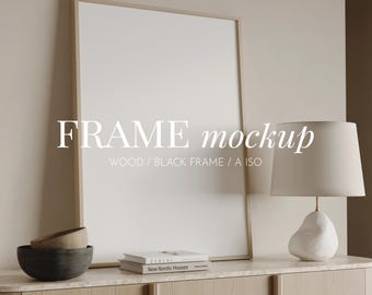 May include: A large, blank picture frame with a light wood border is propped against a neutral wall. The text "FRAME mockup" is displayed on the frame. A lamp with a white shade and a decorative base sits on a wooden cabinet, along with a bowl and books.