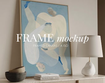 May include: A framed canvas with an abstract painting in shades of blue and white. The artwork features figures in motion. The text "FRAME mockup" is displayed on the canvas. A white lamp and decorative bowls are on a wooden surface.