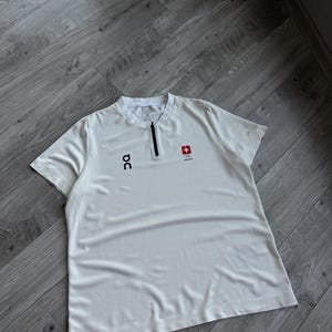 Puede incluir: Camiseta deportiva blanca de manga corta con una pequeña cremallera negra en el cuello. La camiseta presenta el logotipo "on" en negro en el pecho izquierdo y un emblema de la bandera suiza en el derecho. Fabricada con tejido ligero y transpirable.