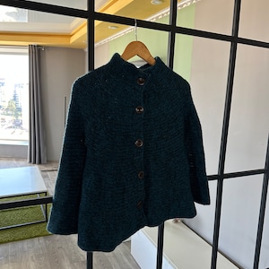May include: A dark teal knitted cardigan with a high neck and button closure. The cardigan is hanging on a wooden hanger.