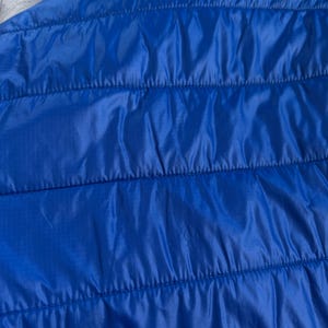 May include: Close-up of a bright blue quilted jacket. The jacket has a hood and horizontal stitching. The fabric has a slight sheen, and the overall appearance is smooth and sleek. The jacket is a vibrant shade of blue.