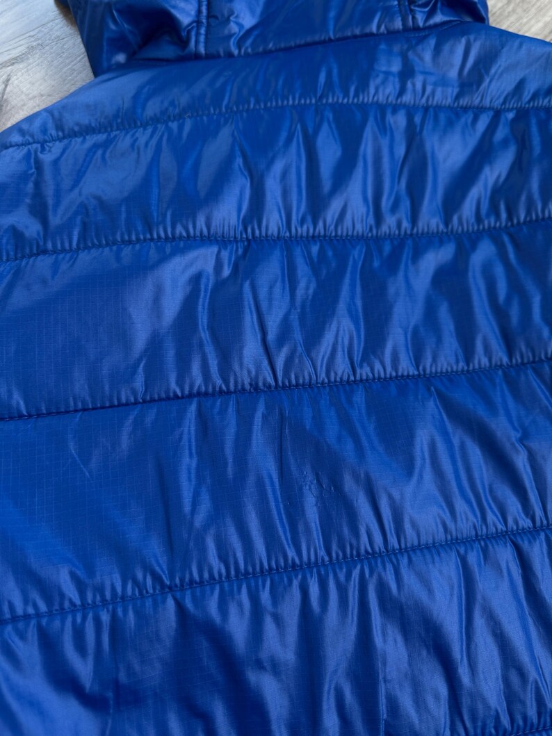 May include: Close-up of a bright blue quilted jacket. The jacket has a hood and horizontal stitching. The fabric has a slight sheen, and the overall appearance is smooth and sleek. The jacket is a vibrant shade of blue.