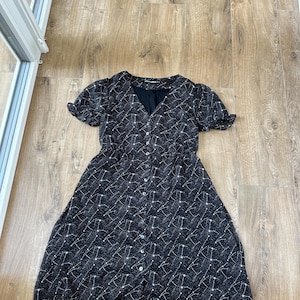 May include: A black dress with a constellation pattern. The dress has short sleeves, a V-neck, and a button-down front. The fabric is a dark color with a white constellation design.