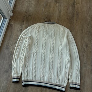 Puede incluir: Su&eacute;ter de punto trenzado color crema con cuello en V y mangas largas. Los pu&ntilde;os y el dobladillo presentan un dise&ntilde;o a rayas en marr&oacute;n, azul marino y crema. El su&eacute;ter est&aacute; hecho de punto grueso y texturizado.