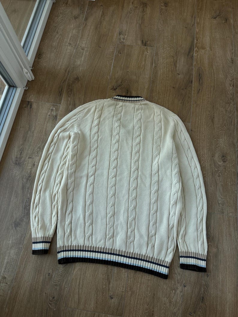 Puede incluir: Su&eacute;ter de punto trenzado color crema con cuello en V y mangas largas. Los pu&ntilde;os y el dobladillo presentan un dise&ntilde;o a rayas en marr&oacute;n, azul marino y crema. El su&eacute;ter est&aacute; hecho de punto grueso y texturizado.