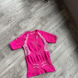Puede incluir: Camiseta deportiva de manga corta rosa con paneles de malla blanca y el texto "X-BIONIC" en el lado izquierdo y "INNOVATION" en el lado derecho.