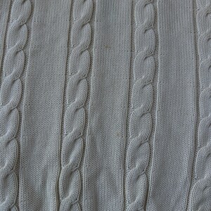 Puede incluir: Primer plano de una tela de punto beige claro con patrones de punto de cable verticales. La tela tiene un aspecto texturizado con secciones alternas de cable y punto liso. El borde inferior presenta un borde acanalado marr&oacute;n m&aacute;s oscuro.