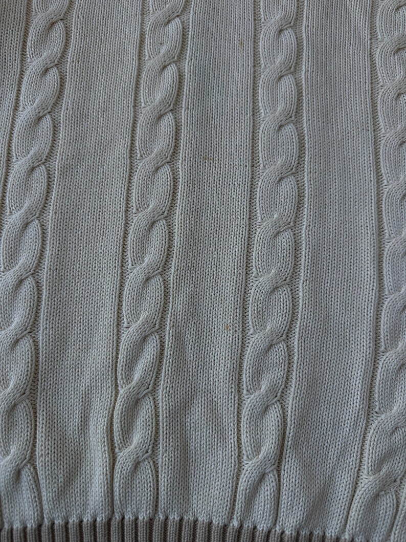 Puede incluir: Primer plano de una tela de punto beige claro con patrones de punto de cable verticales. La tela tiene un aspecto texturizado con secciones alternas de cable y punto liso. El borde inferior presenta un borde acanalado marr&oacute;n m&aacute;s oscuro.