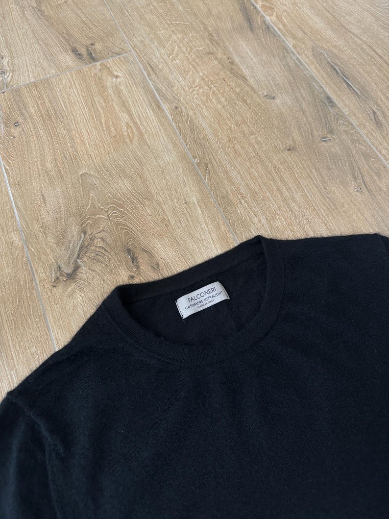 Puede incluir: Un su&eacute;ter negro de manga larga con cuello redondo. Una etiqueta rectangular blanca est&aacute; cosida en el interior del cuello, con el texto FALCONERI, CASHMERE ULTRALIGHT y MADE IN ITALY en letras negras. La prenda tiene una textura de punto fino y ligero.