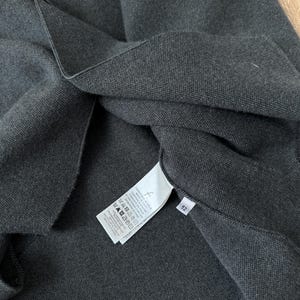 Op de afbeelding: Een donker antracietgrijs gebreid kledingstuk met een wit waslabel aan de binnenzijde met gedrukte tekst en wassymbolen, naast een klein wit label met het nummer 52. De stof heeft een zichtbaar, fijn geweven textuur over het hele oppervlak.