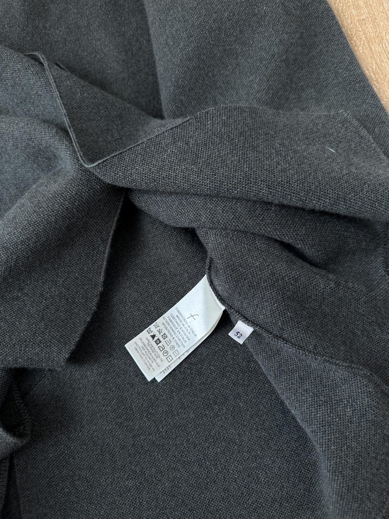 Op de afbeelding: Een donker antracietgrijs gebreid kledingstuk met een wit waslabel aan de binnenzijde met gedrukte tekst en wassymbolen, naast een klein wit label met het nummer 52. De stof heeft een zichtbaar, fijn geweven textuur over het hele oppervlak.