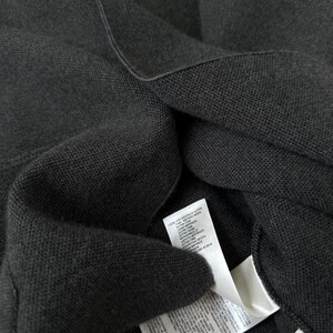 Op de afbeelding: Een donker antracietgrijs gebreid kledingstuk met een fijne textuur. Twee witte onderhouds- en materiaallabels zijn aan de binnenkant bevestigd, met tekst in meerdere talen waaronder 100% extra fijne merinowol, en een klein label met maat 52.