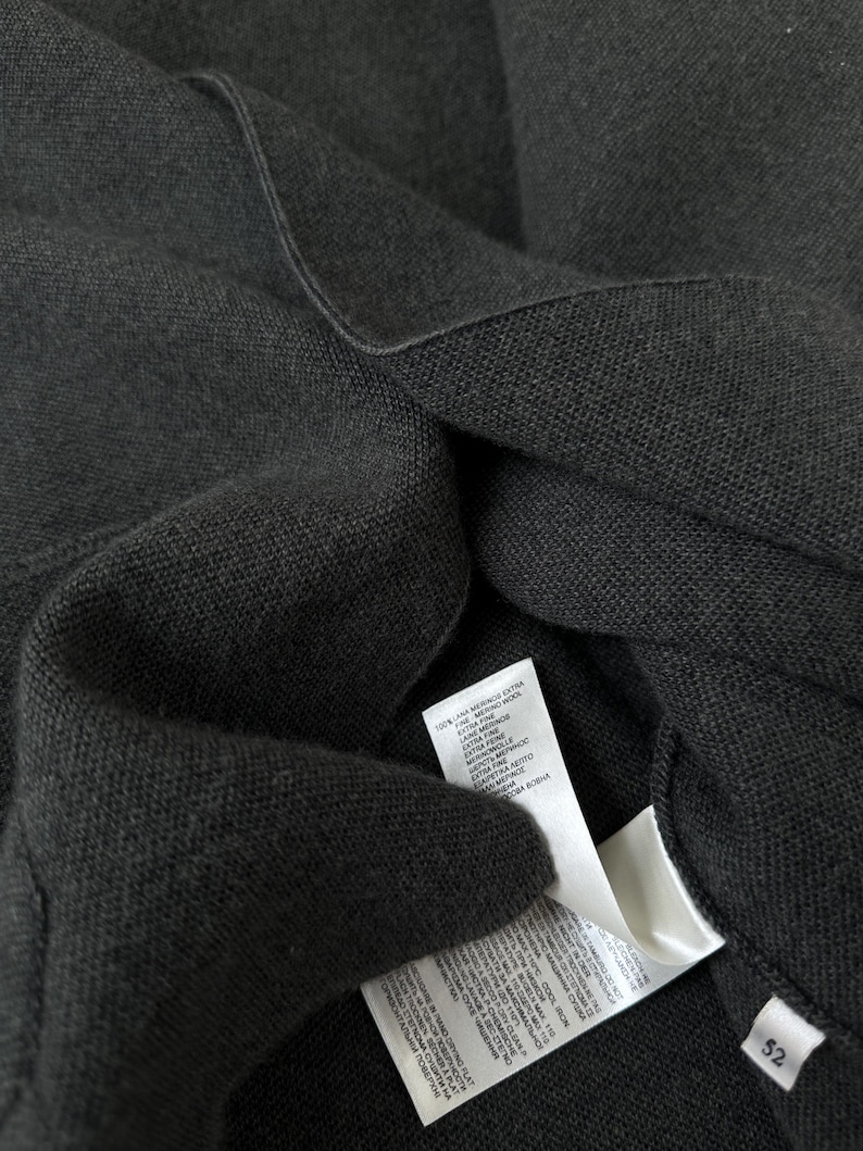 Op de afbeelding: Een donker antracietgrijs gebreid kledingstuk met een fijne textuur. Twee witte onderhouds- en materiaallabels zijn aan de binnenkant bevestigd, met tekst in meerdere talen waaronder 100% extra fijne merinowol, en een klein label met maat 52.