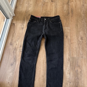 May include: A pair of black denim jeans featuring a button fly closure, five pockets, and a small pink brand label attached to the interior waistband. The jeans have a straight-leg cut with visible stitching along the seams and a classic denim texture.