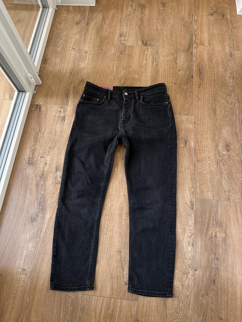 May include: A pair of black denim jeans featuring a button fly closure, five pockets, and a small pink brand label attached to the interior waistband. The jeans have a straight-leg cut with visible stitching along the seams and a classic denim texture.