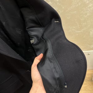 May include: A dark navy blue wool coat with a tailored fit. The interior lining is black and features a logo. The coat has a classic design with a notched lapel and a single-breasted closure. The coat is made of a thick, warm material.