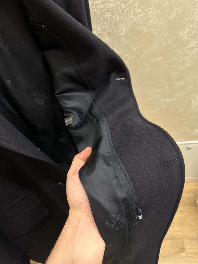 May include: A dark navy blue wool coat with a tailored fit. The interior lining is black and features a logo. The coat has a classic design with a notched lapel and a single-breasted closure. The coat is made of a thick, warm material.