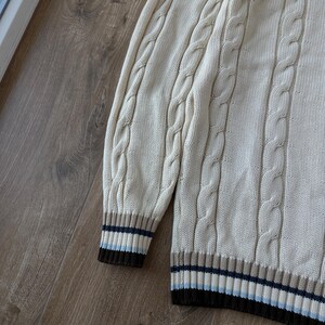 Puede incluir: Su&eacute;ter de punto trenzado color crema con dobladillo y pu&ntilde;os acanalados. Los pu&ntilde;os y el dobladillo presentan rayas marrones, azules y beige. El su&eacute;ter tiene un dise&ntilde;o cl&aacute;sico y texturizado.