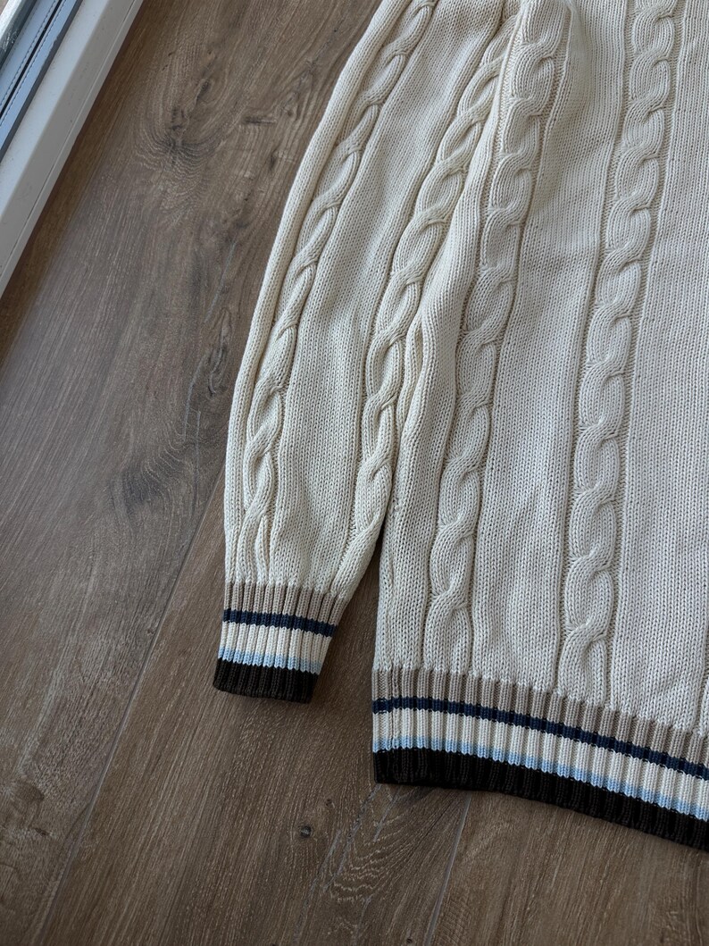 Puede incluir: Su&eacute;ter de punto trenzado color crema con dobladillo y pu&ntilde;os acanalados. Los pu&ntilde;os y el dobladillo presentan rayas marrones, azules y beige. El su&eacute;ter tiene un dise&ntilde;o cl&aacute;sico y texturizado.