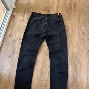 May include: A pair of black denim jeans shown from the back, featuring two rear pockets, a waistband with belt loops, and a dark rectangular leather-like patch on the right side of the waistband with a small red tag attached to the corner.