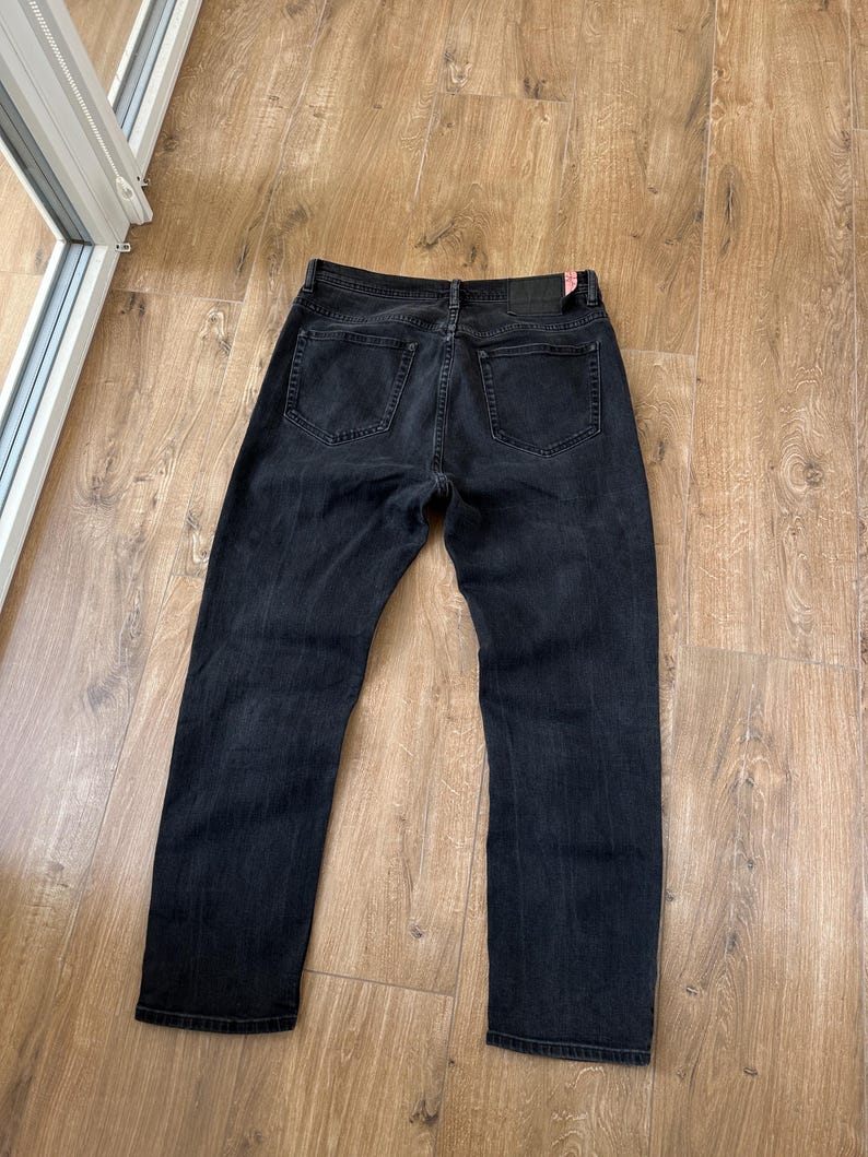 May include: A pair of black denim jeans shown from the back, featuring two rear pockets, a waistband with belt loops, and a dark rectangular leather-like patch on the right side of the waistband with a small red tag attached to the corner.