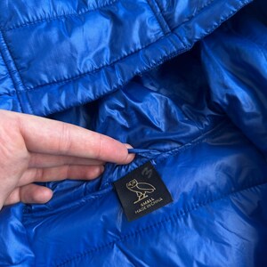 May include: A close-up of a bright blue quilted jacket. The jacket has a hood and a small black tag with a logo and the text "SMALL MADE IN CHINA". The jacket is made of a shiny, lightweight material.