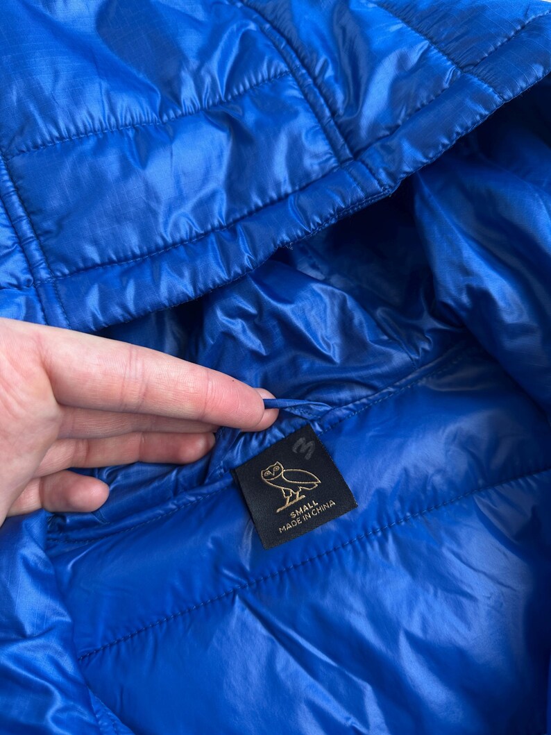 May include: A close-up of a bright blue quilted jacket. The jacket has a hood and a small black tag with a logo and the text "SMALL MADE IN CHINA". The jacket is made of a shiny, lightweight material.