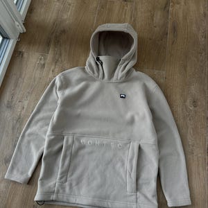 Puede incluir: Una sudadera con capucha gris claro con cuello alto y bolsillo tipo canguro. La palabra "MONTEC" está bordada en el bolsillo. La sudadera tiene un pequeño logotipo en el pecho y un cordón en la capucha.