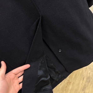 May include: A close-up of a black blazer, showcasing the fabric's texture and tailoring details. The interior lining is visible, with a button closure and a vent at the back. The blazer appears to be made of a wool blend.