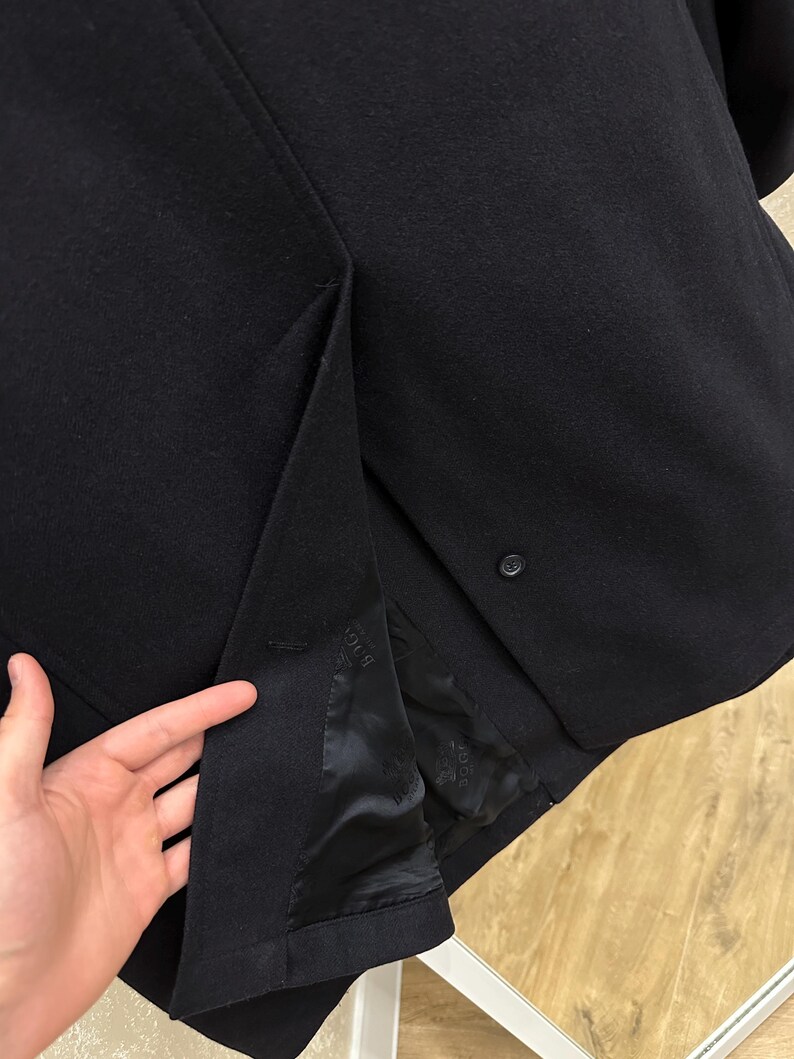 May include: A close-up of a black blazer, showcasing the fabric's texture and tailoring details. The interior lining is visible, with a button closure and a vent at the back. The blazer appears to be made of a wool blend.
