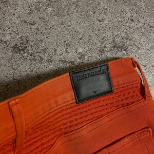 May include: Orange jeans with a black leather patch that says "True Religion" on the back waistband.