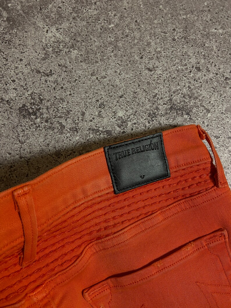 May include: Orange jeans with a black leather patch that says "True Religion" on the back waistband.