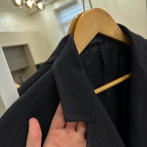 May include: A black blazer hanging on a wooden hanger. The blazer has a notched lapel and a classic design. The image is taken indoors, with a mirror and lights in the background.