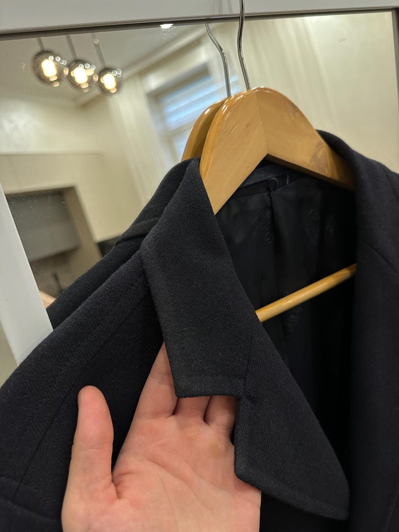 May include: A black blazer hanging on a wooden hanger. The blazer has a notched lapel and a classic design. The image is taken indoors, with a mirror and lights in the background.