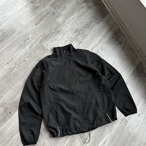 Peut inclure: Veste noire avec fermeture éclair intégrale, col montant et deux poches latérales. La veste a un cordon de serrage en bas et une bordure blanche le long des poches et de la fermeture éclair. La veste est posée sur un sol en bois.