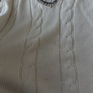 Puede incluir: Su&eacute;ter de punto trenzado color crema con cuello en V. El cuello presenta un dise&ntilde;o a rayas en azul marino, marr&oacute;n y crema. El su&eacute;ter tiene un patr&oacute;n de punto trenzado cl&aacute;sico y est&aacute; hecho de un material suave y texturizado.