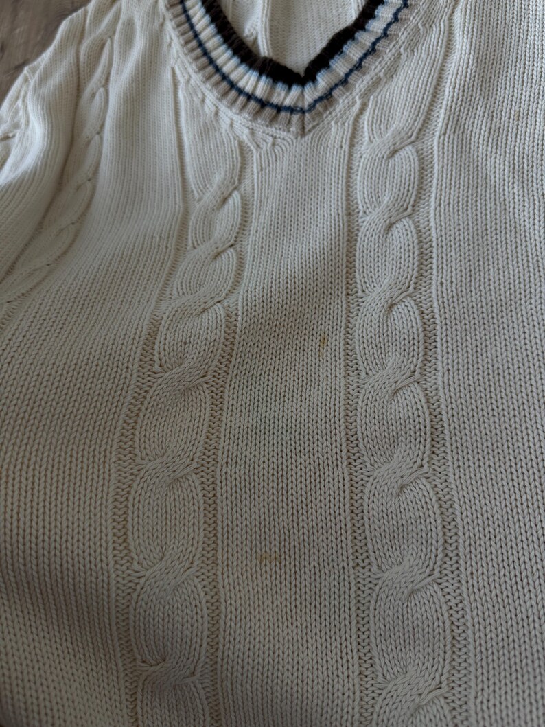 Puede incluir: Su&eacute;ter de punto trenzado color crema con cuello en V. El cuello presenta un dise&ntilde;o a rayas en azul marino, marr&oacute;n y crema. El su&eacute;ter tiene un patr&oacute;n de punto trenzado cl&aacute;sico y est&aacute; hecho de un material suave y texturizado.