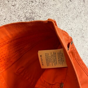 May include: Close-up of the inside of a pair of orange denim pants. The fabric content label is visible and reads: "Made in USA, In den USA hergestellt, Fabrique aux E-U., Hecho en EE.UU., Self/Eigenstoff/Corps de la Misma Tela, 92% Cotton/Baumwolle/Coton/Algod&oacute;n, 8% ElastereLL-P, 2% Elastane/Elastan/Elastahanne/Elastano"