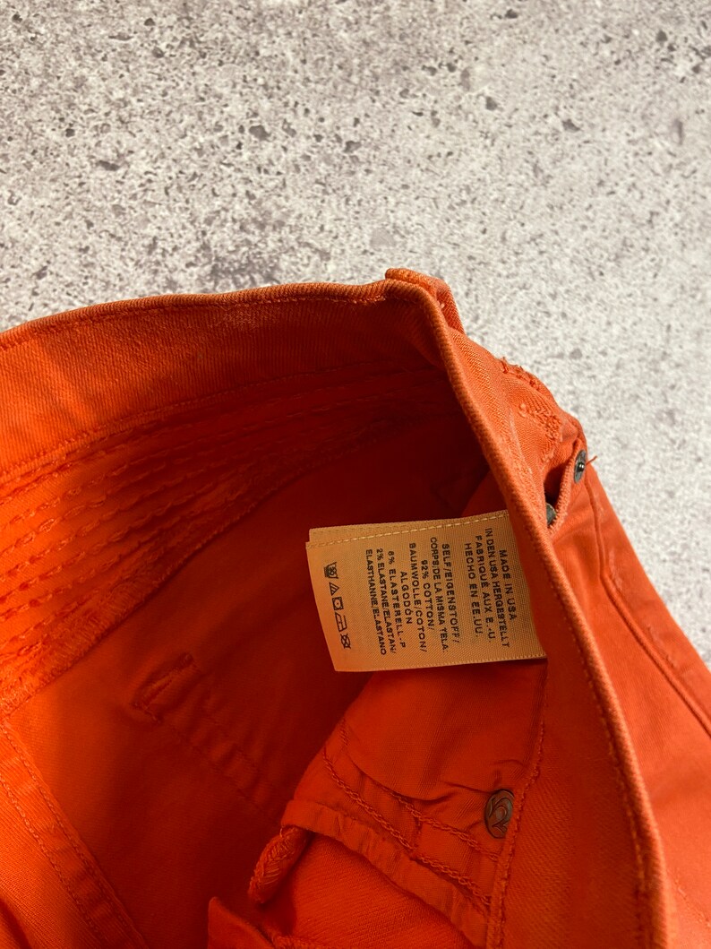 May include: Close-up of the inside of a pair of orange denim pants. The fabric content label is visible and reads: "Made in USA, In den USA hergestellt, Fabrique aux E-U., Hecho en EE.UU., Self/Eigenstoff/Corps de la Misma Tela, 92% Cotton/Baumwolle/Coton/Algod&oacute;n, 8% ElastereLL-P, 2% Elastane/Elastan/Elastahanne/Elastano"