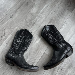May include: A pair of black leather cowboy boots with detailed stitching. The boots feature pointed toes and stacked heels. The leather has a shiny finish, and they are placed on a light grey wooden floor.
