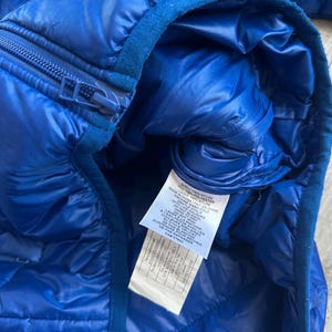 May include: Close-up of a vibrant blue quilted jacket. The jacket features a zipper closure and a soft, dark blue collar. A tag with care instructions is visible. The jacket is made of nylon and polyester.
