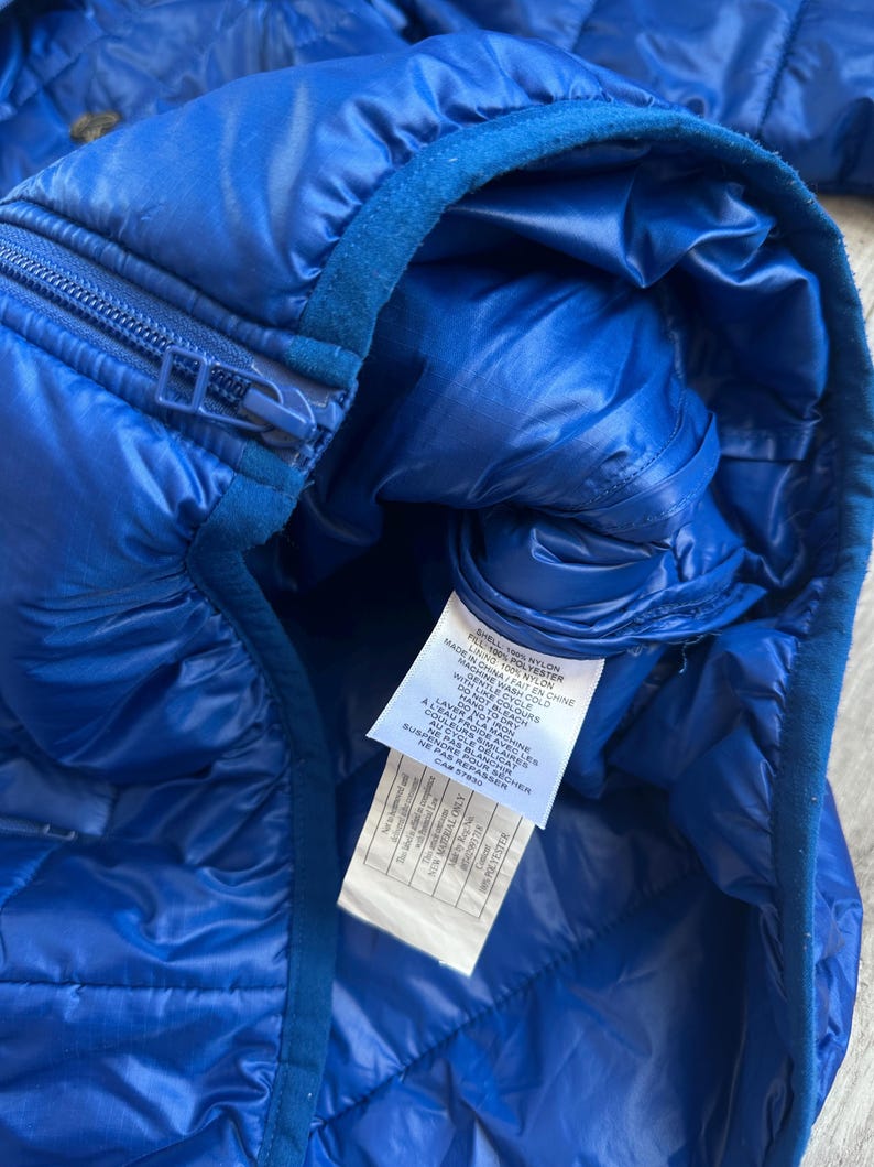 May include: Close-up of a vibrant blue quilted jacket. The jacket features a zipper closure and a soft, dark blue collar. A tag with care instructions is visible. The jacket is made of nylon and polyester.